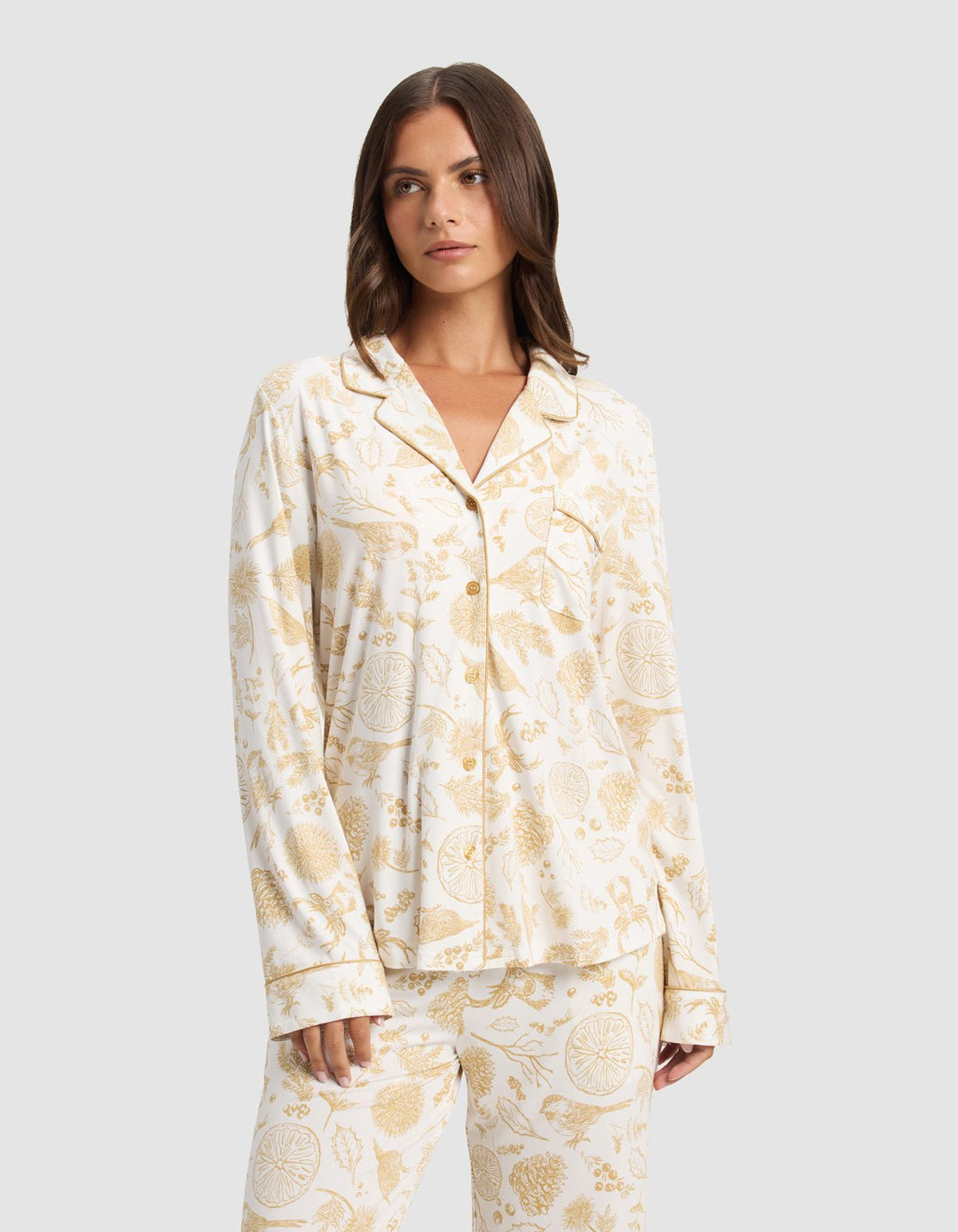 A woman wears the Cozy Earth Women's Bamboo Stretch-Knit Long Sleeve Pajama Top with a white base and light brown botanical and animal print, standing against a plain light gray background.|Color:Winter Toile Dijon