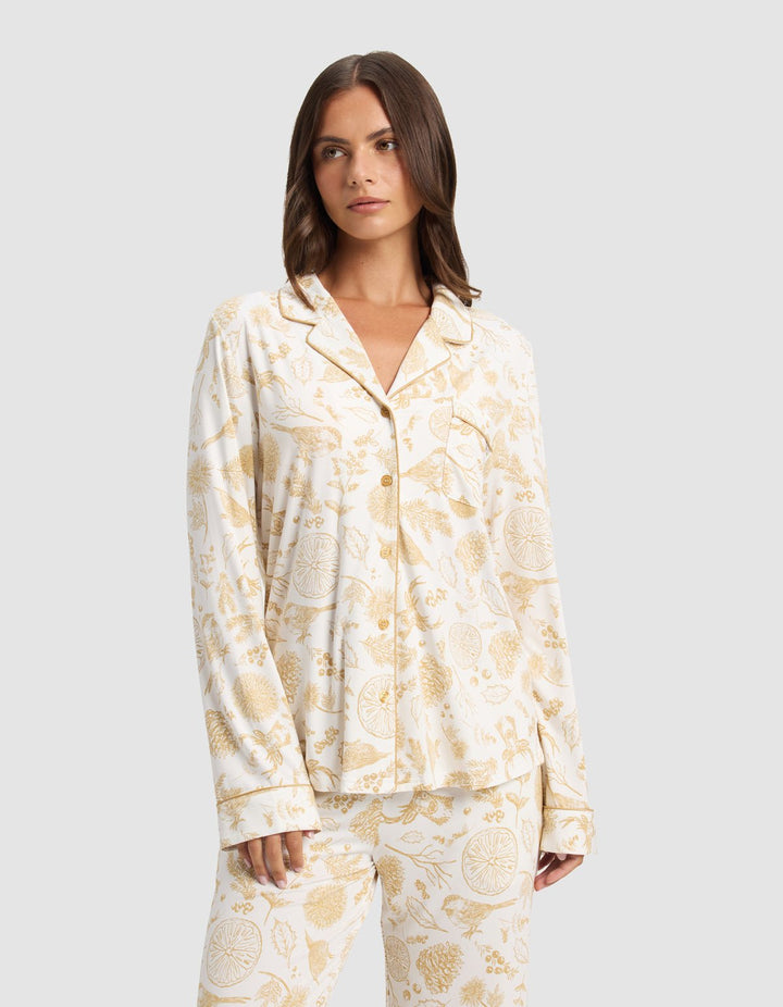 A woman wears the Cozy Earth Women's Bamboo Stretch-Knit Long Sleeve Pajama Top with a white base and light brown botanical and animal print, standing against a plain light gray background.|Color:Winter Toile Dijon