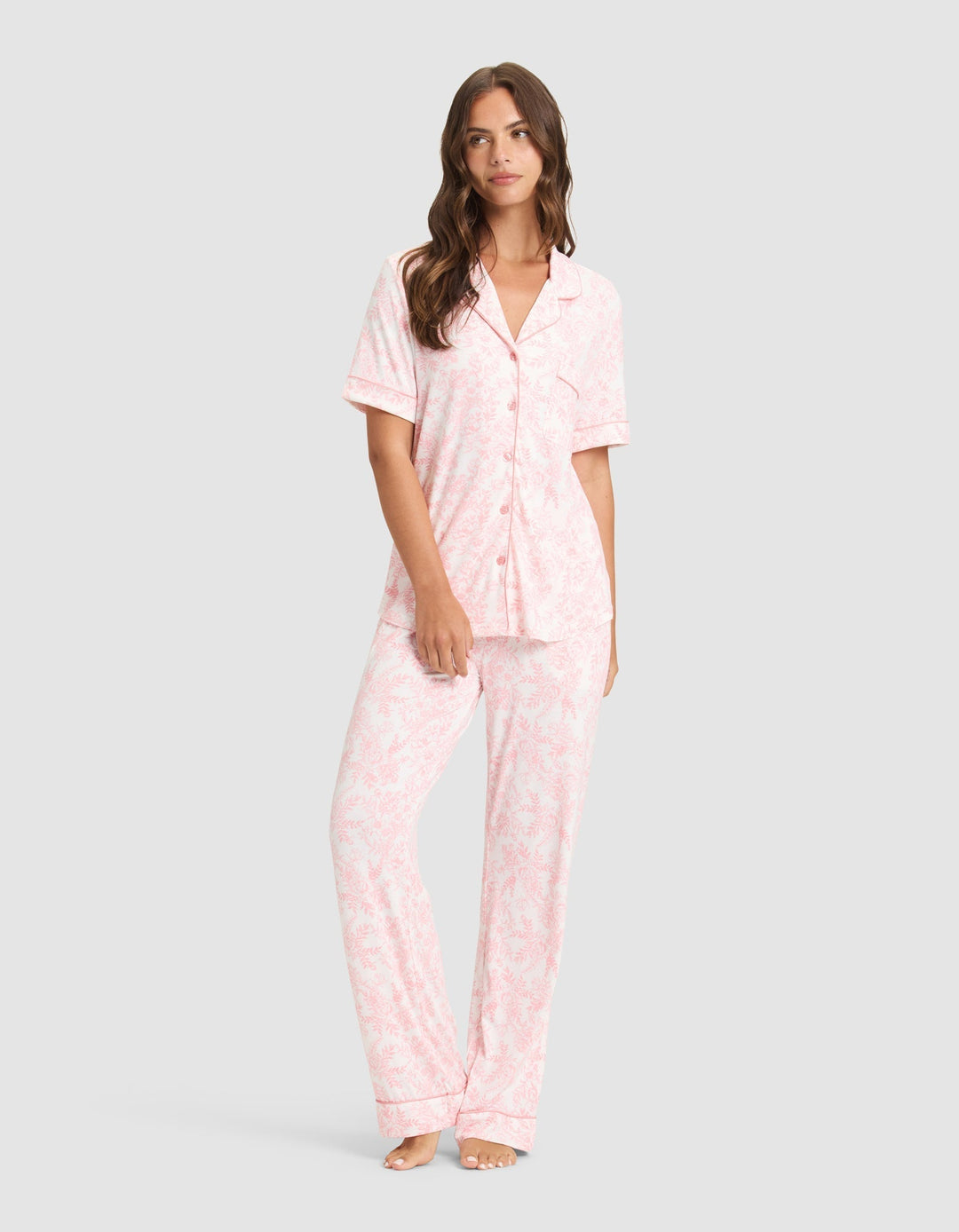 A woman stands barefoot in Cozy Earth's Women's Bamboo Stretch-Knit Long Sleeve Pajama Set, featuring light pink, long pants and a matching button-up top with a subtle floral pattern, against a light gray background. |Color:Tulip Toile