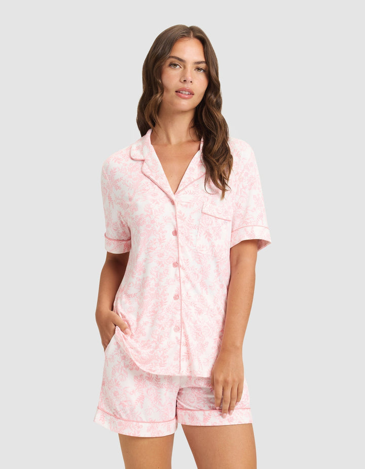 A woman with long brown hair models the HIDE Women's Bamboo Stretch-Knit Short Sleeve Pajama Top in light pink, featuring a subtle floral pattern, button-up front, and matching shorts, standing against a plain light background. |Color:Tulip Toile