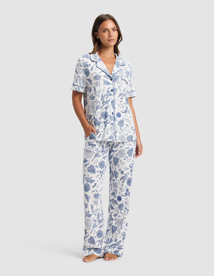 A woman stands barefoot in the Cozy Earth Women's Bamboo Stretch-Knit Short Sleeve Pajama Set, featuring a blue and white botanical print with a collared button-up top and loose pants. She has one hand in her pocket and looks relaxed.|Color:Winter Toile Pacific