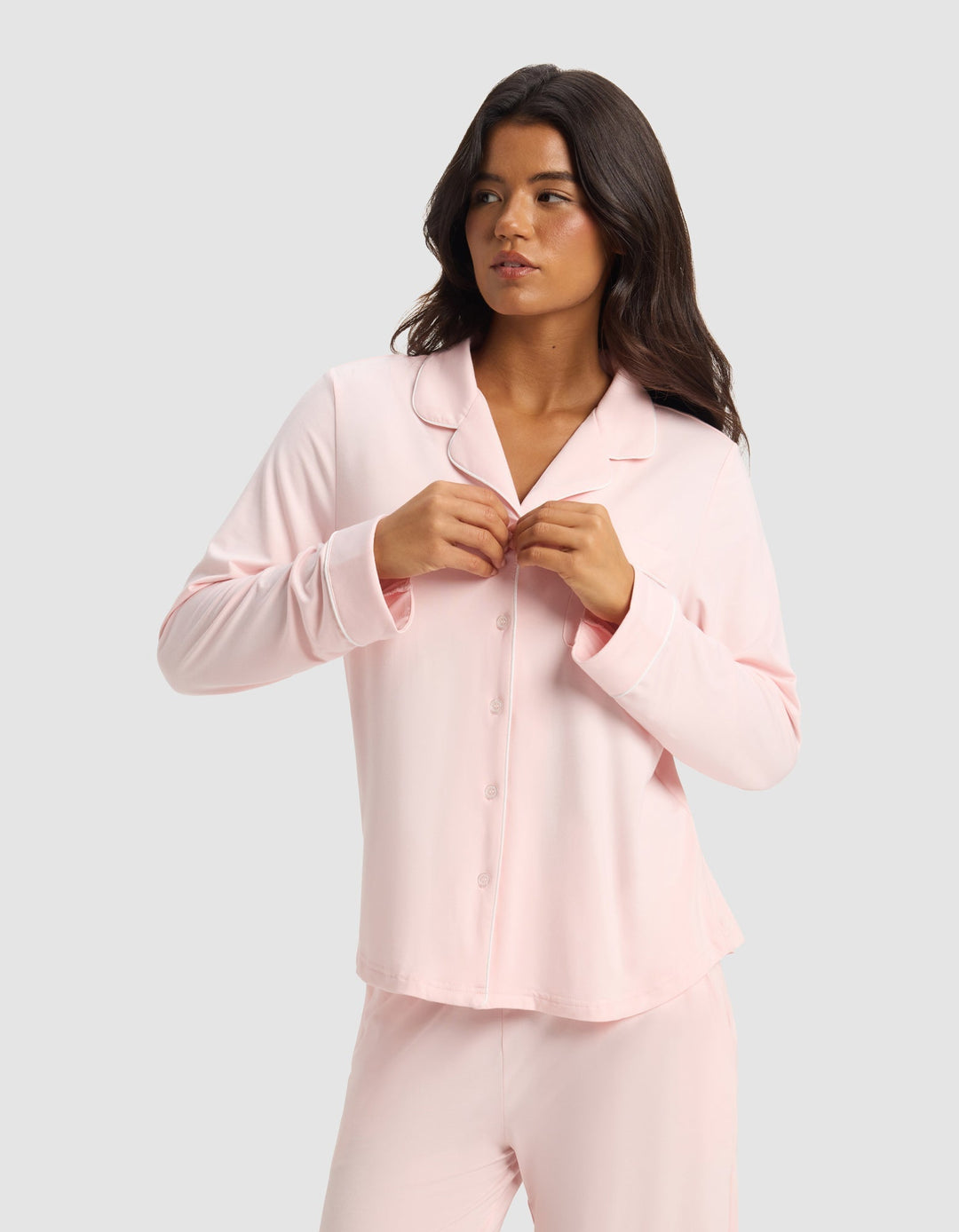 A woman with long dark hair wears the Cozy Earth Women's Bamboo Stretch-Knit Long Sleeve Pajama Set in light pink, fastening a button on her top as she stands against a plain light gray background, looking slightly to her left.|Color:Blossom