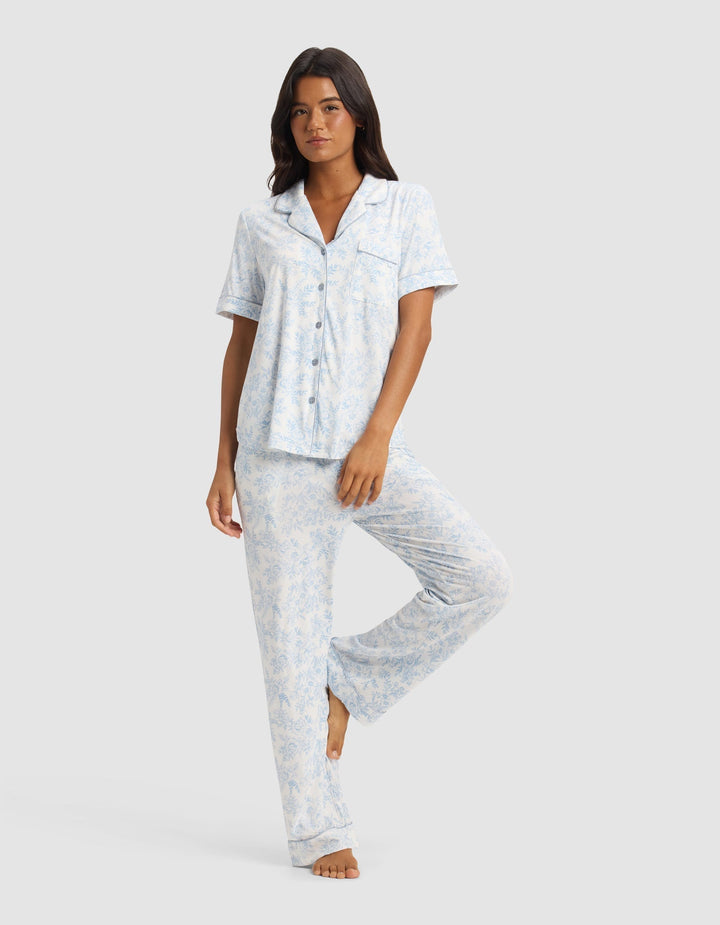 A woman poses barefoot against a plain light background, wearing the Cozy Earth Women's Bamboo Stretch-Knit Short Sleeve & Pant Pajama Set in a light blue and white pattern.|Color:Sky Toile