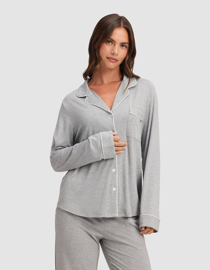 A woman in the HIDE Women's Bamboo Stretch-Knit Long Sleeve Pajama Top, light gray with white piping and buttons, stands against a plain background, softly smiling as she gently holds the top near her chest. |Color:Grey