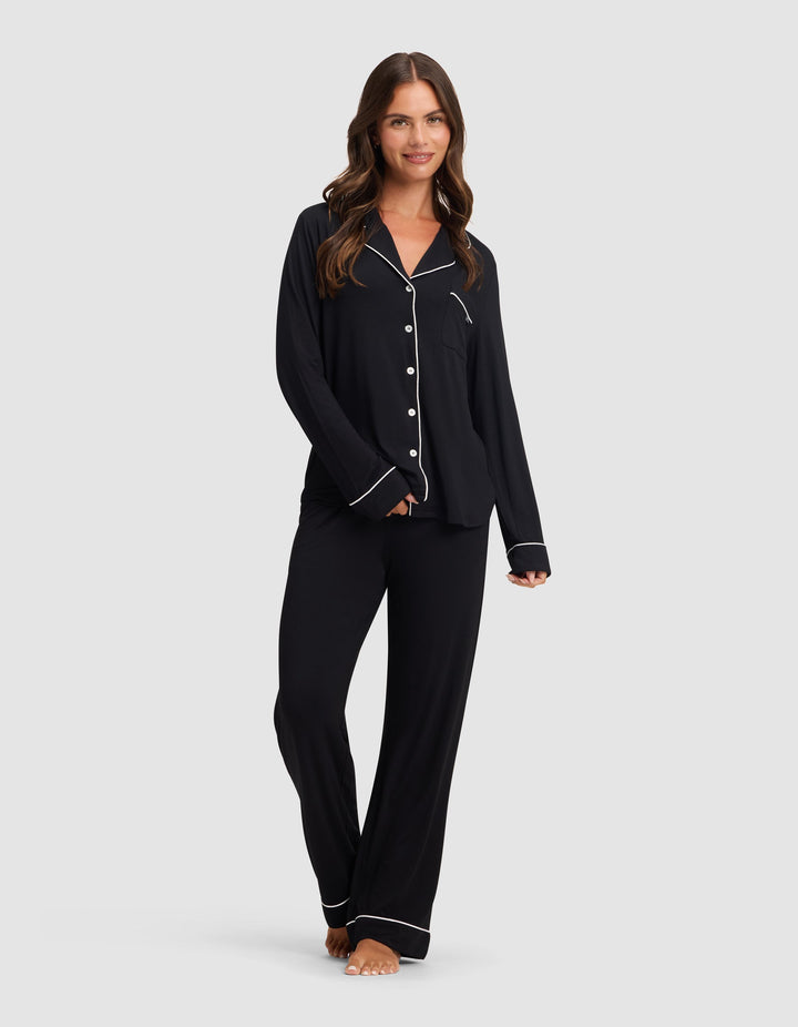 A woman with long brown hair, barefoot and smiling, models the Cozy Earth Women's Bamboo Stretch-Knit Pajama Pant in black with white piping, paired with a matching button-up top, against a plain light gray background.|Color:Black