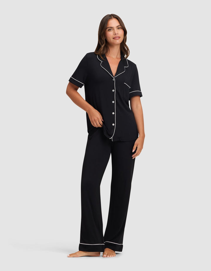 A woman stands barefoot against a light gray background, wearing Cozy Earth’s Women's Bamboo Stretch-Knit Short Sleeve & Pant Pajama Set in black with white piping. The set includes a short-sleeved button-up top and long pants. |Color:Black