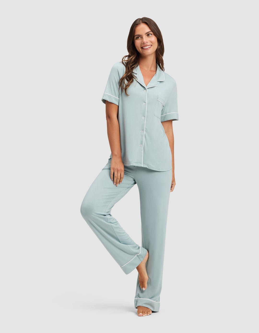 A woman with long brown hair smiles while wearing the Cozy Earth Women's Bamboo Stretch-Knit Short Sleeve & Pant Pajama Set, standing barefoot against a plain white background. |Color:Brook