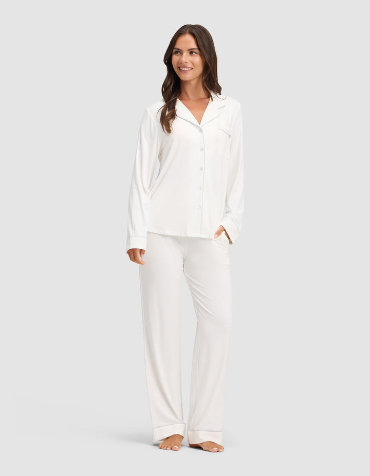 A woman smiles while wearing the Cozy Earth Women's Bamboo Stretch-Knit Long Sleeve Pajama Set, featuring a button-up top. She stands barefoot against a plain light gray background. |Color:Ivory