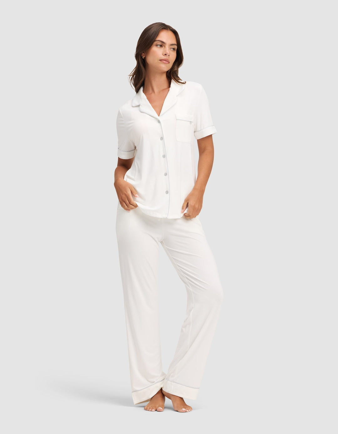 A woman wears Cozy Earth's Women's Bamboo Stretch-Knit Short Sleeve & Pant Pajama Set, featuring a white short-sleeved button-up top with chest pocket and matching long pants, standing barefoot against a plain light gray background. |Color:Ivory
