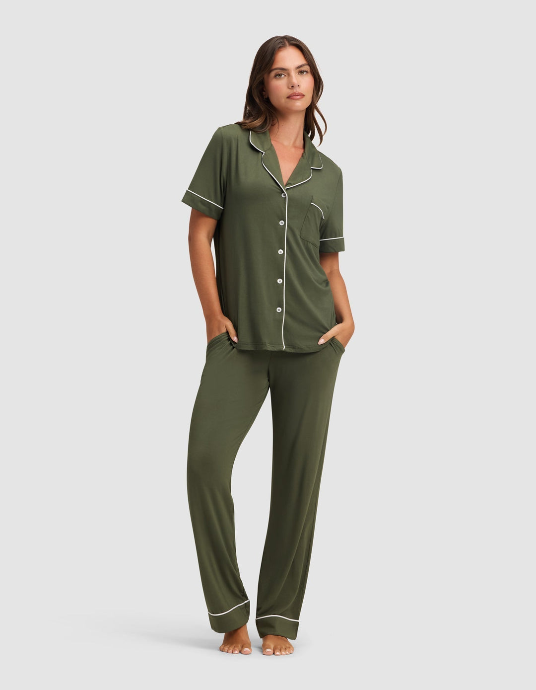 A woman stands barefoot in the Cozy Earth Women's Bamboo Stretch-Knit Short Sleeve & Pant Pajama Set in olive green with white piping, featuring a button-up top and long pants, her hands in her pockets against a plain light background.|Color:Olive