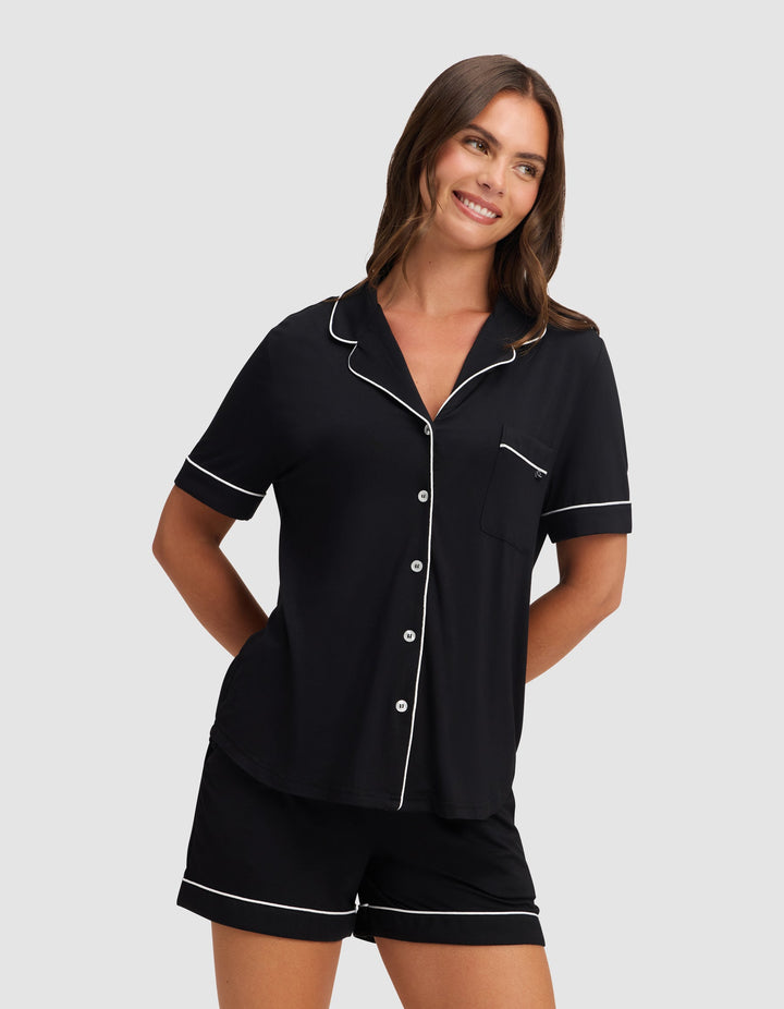 A woman smiles wearing HIDE's Women's Bamboo Stretch-Knit Short Sleeve Pajama Top in black with white piping, featuring a button-up front and chest pocket, paired with matching shorts against a plain light gray background. |Color:Black