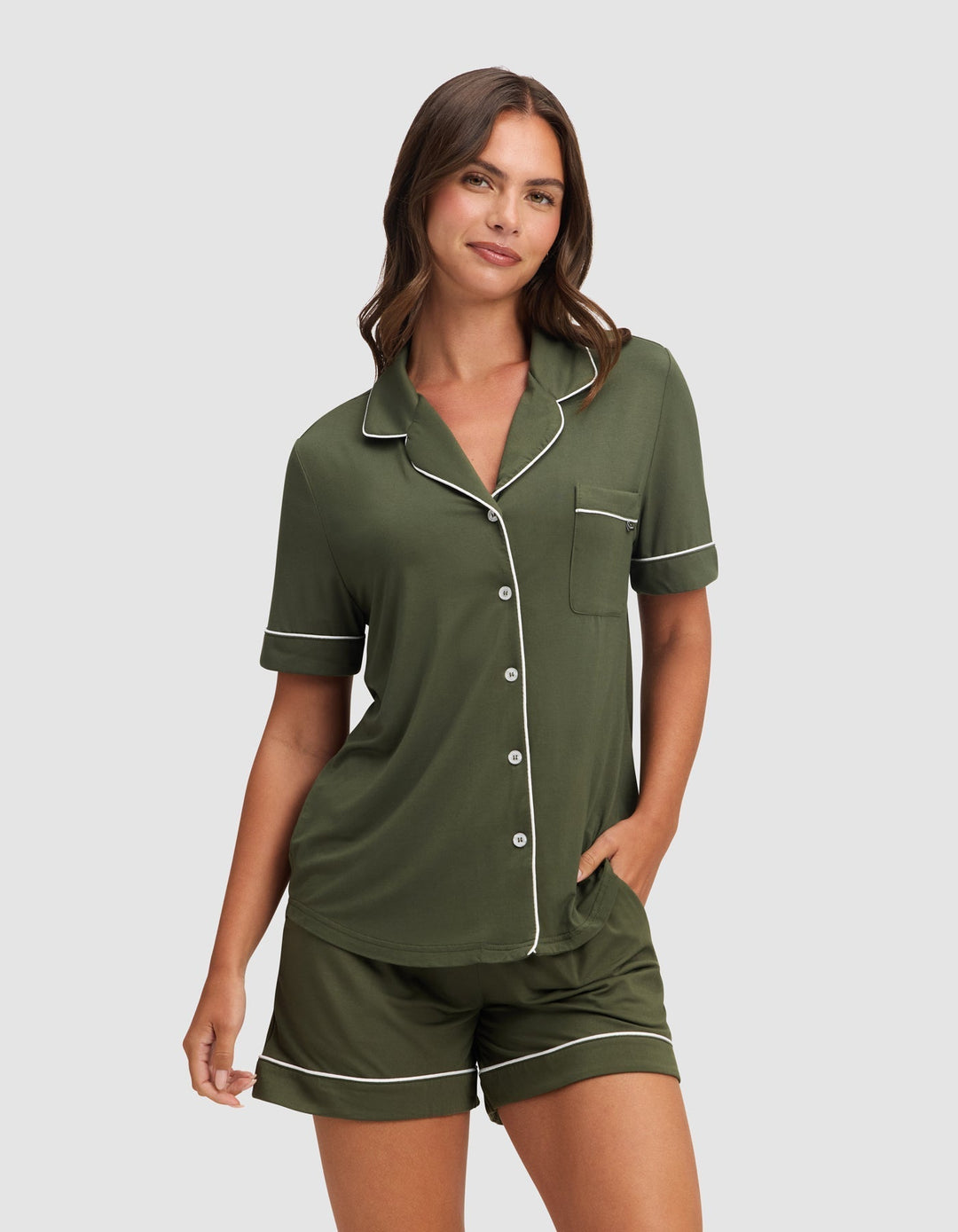 A woman stands against a plain background wearing the HIDE Women's Bamboo Stretch-Knit Short Sleeve Pajama Top in Olive, featuring white piping, front buttons, a chest pocket, and matching shorts with one hand in her pocket. |Color:Olive